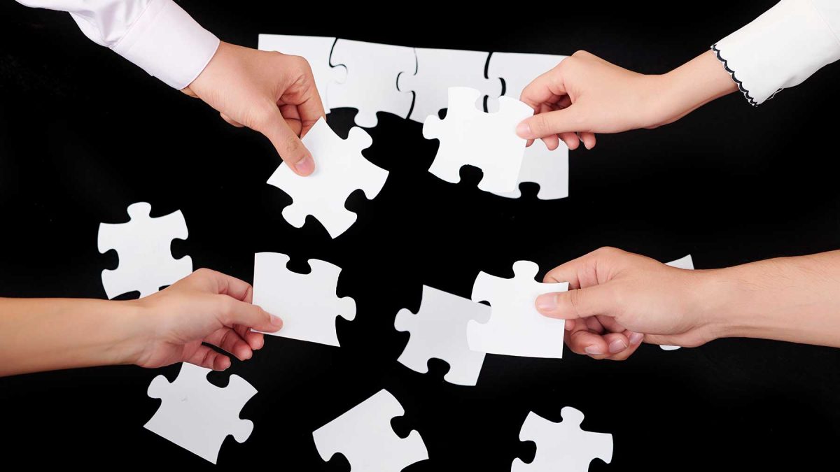 Top down view of four people's hands placing white jigsaw pieces into a puzzle.